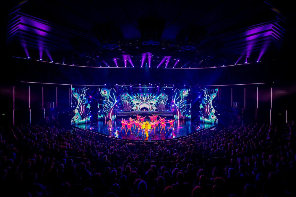 Tänzerinnen in rosa Federkostümen und eine Person im gelben Federkostüm performen vor farbenreichen Pfauenfeder Projektionen im voll besetzten Friedrichstadt Palast.