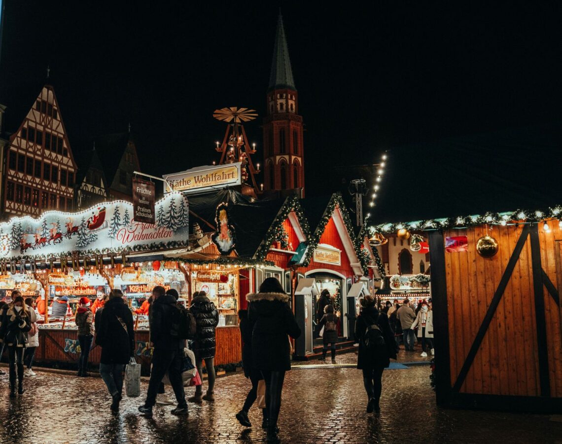 Belebter Weihnachtsmarkt bei Nacht mit festlich beleuchteten Ständen, Menschen und einer Kirche im Hintergrund