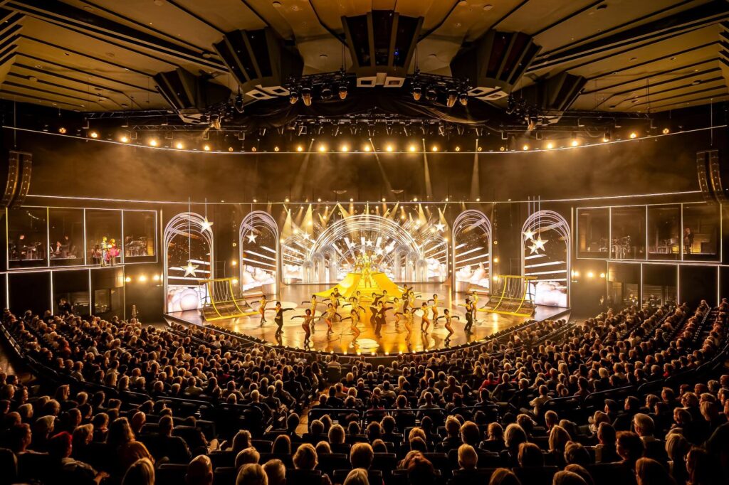 Große Theaterbühne mit vielen tanzenden Darstellenden, heller Showbeleuchtung und voll besetztem Zuschauerraum.