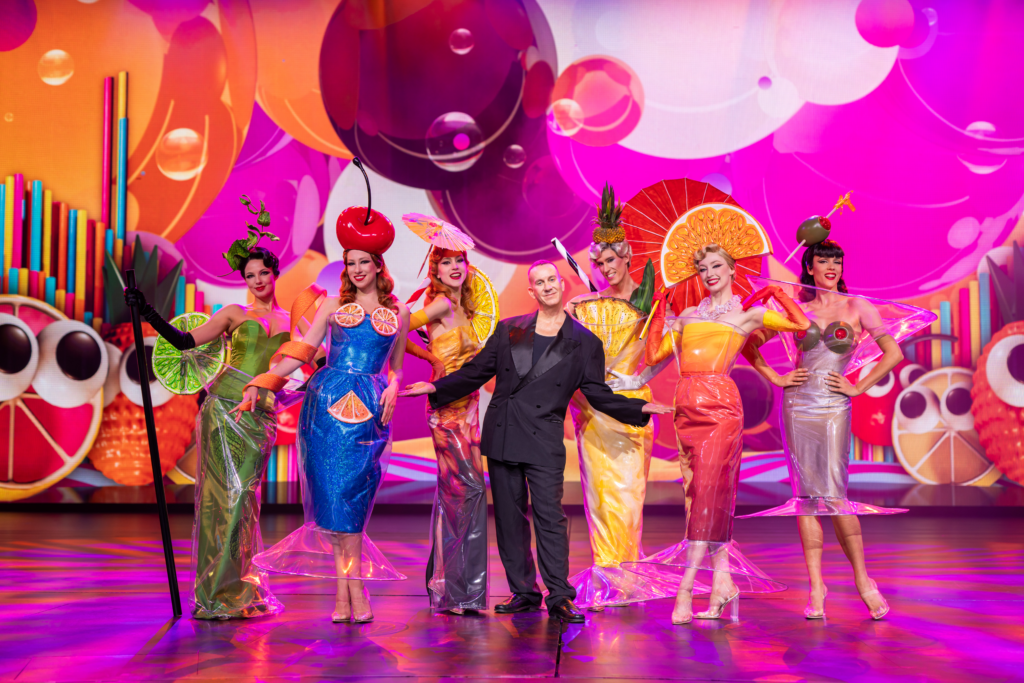 Eine Gruppe von Personen steht auf einer Bühne vor einer farbenfrohen, fruchtinspirierten Kulisse, alle in auffälligen, kostümhaften Outfits gestaltet wie verschiedene Obstsorten. In der Mitte steht eine Person in dunkler Kleidung, umgeben von den kostümierten Darsteller*innen.