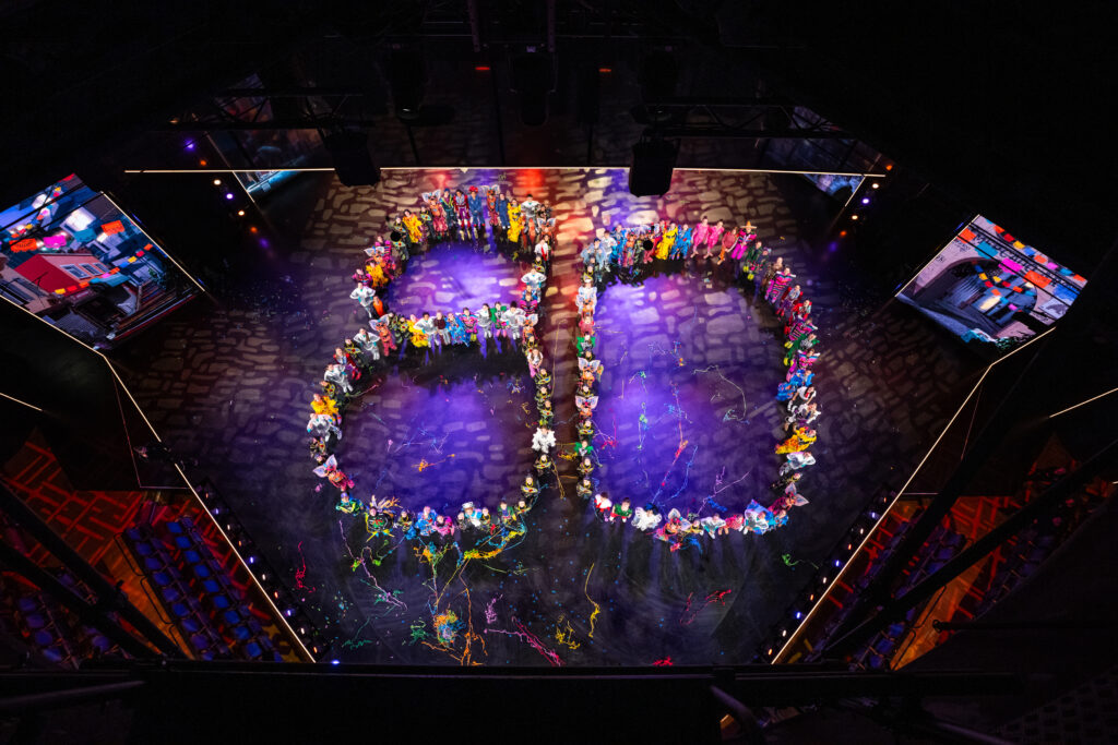 Kinder und Jugendliche auf einer Bühne bilden die Zahl 80 in bunten Kostümen.