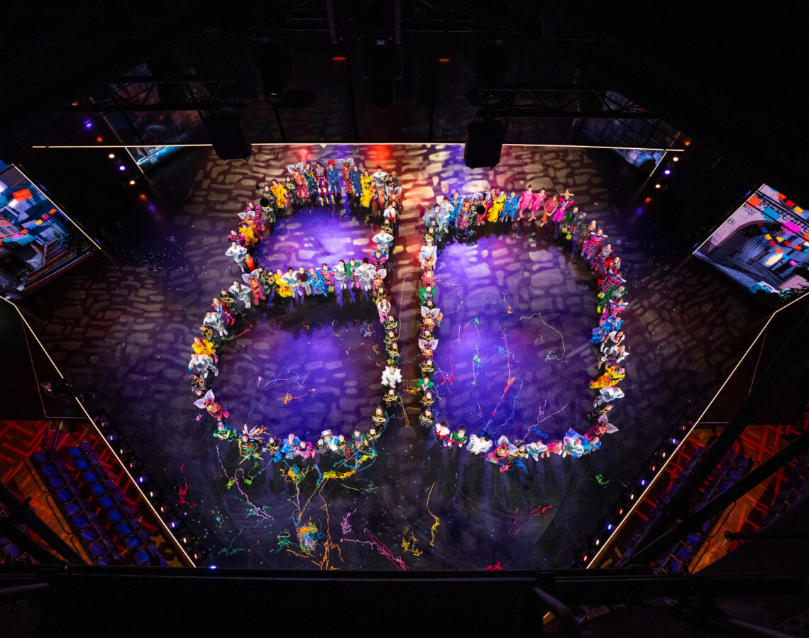 Kinder und Jugendliche auf einer Bühne bilden die Zahl 80 in bunten Kostümen.