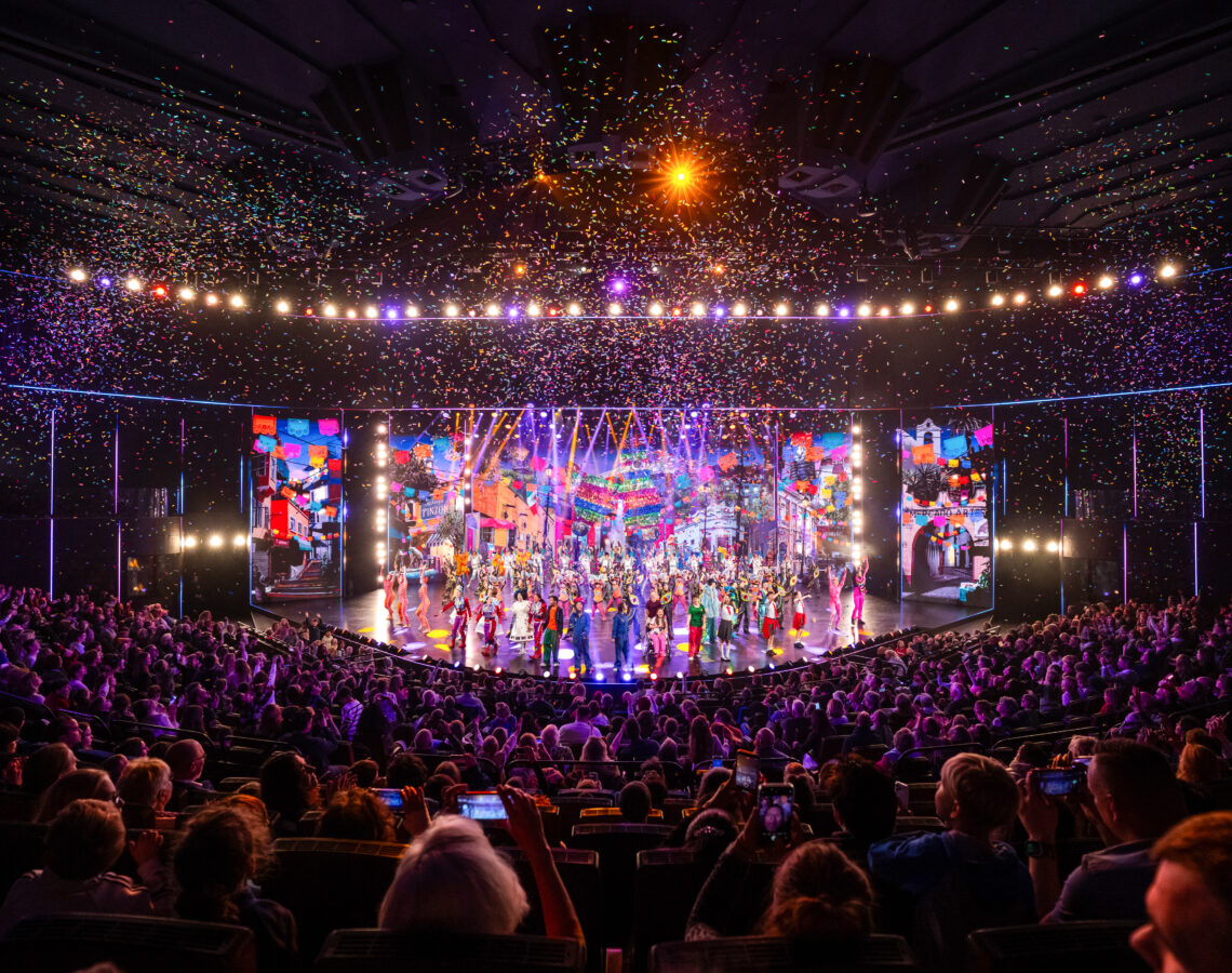 bunte Bühne mit Kindern im Zentrum. Im Vordergrund ein klatschendes Publikum