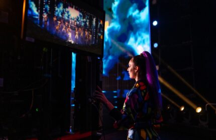 Seitliche Aufnahme einer Performerin backstage, die eine Show auf einem Monitor verfolgt.