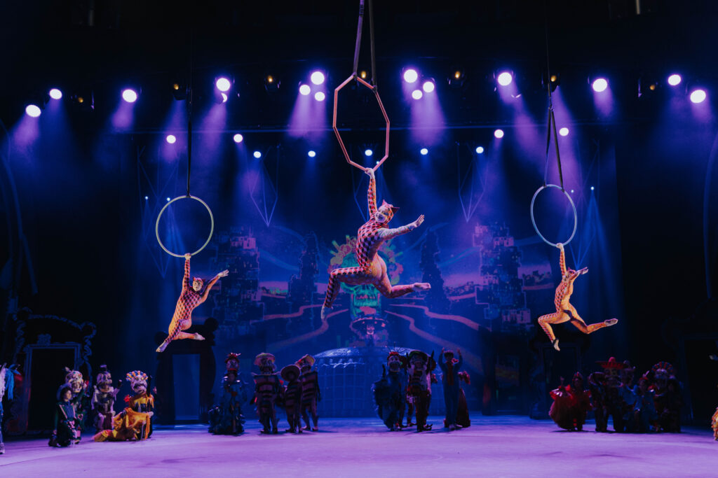 Drei Artistinnen führen eine akrobatische Luftnummer an Ringen aus, vor einer bunten, leuchtenden Bühne 