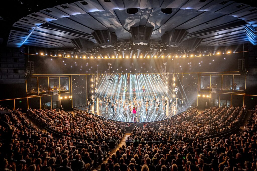 Große Theaterbühne mit vielen Tänzerinnen und Tänzern unter hellem Bühnenlicht, davor ein voll besetzter Saal.