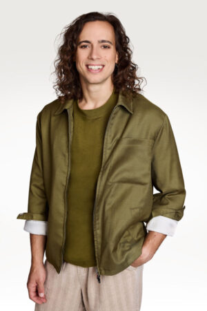 Porträt von Antonio Calanna, mit dunkelbraunem, lockigem Haar, olivgrünem T-Shirt und grüner offener Jacke sowie heller Hose vor neutralem Studiohintergrund.